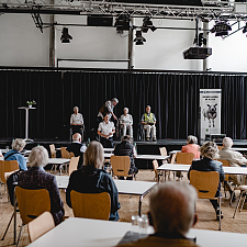 Sitzende ältere Personen vor einer Bühne auf der eine Polizistin steht