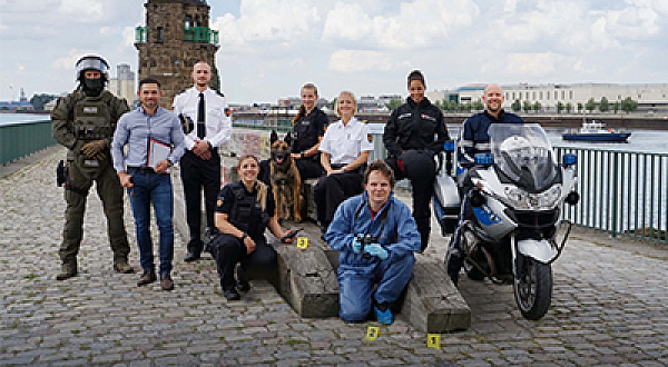 Auf dem Foto sind ein Motorrad sowie neun Personen zu sehen, die die unterschiedlichen Uniformen der Polizei tragen