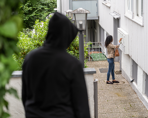 Vermummte Person, die aus der Entfernung eine Frau am Postkasten eines Hauses beobachtet